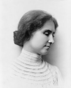 A black and white portrait photograph of Helen Keller. She is shown in profile view, facing to the right with a serene expression. Her eyes are closed, and her hair is styled in a neat, low bun. She is wearing a high-necked, lace-trimmed blouse with intricate detailing