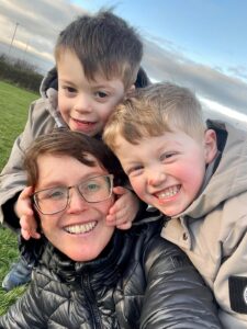 Nia with brown hair, glasses, and a bright smile is outdoors on a grassy field with her two boys. They are both wearing light grey jackets, are playfully embracing her, with one gently holding her face. They all have joyful expressions. The background features a blue sky with scattered clouds.