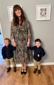 Nia with long brown hair and a warm smile, holding hands with her two young boys. She is wearing a flowing leopard-print dress and black heels. The boys, both dressed in matching navy blue polka-dot shirts, beige trousers, and brown boots, grin cheerfully. They are at home and the background features a neutral-coloured wall with framed photographs.